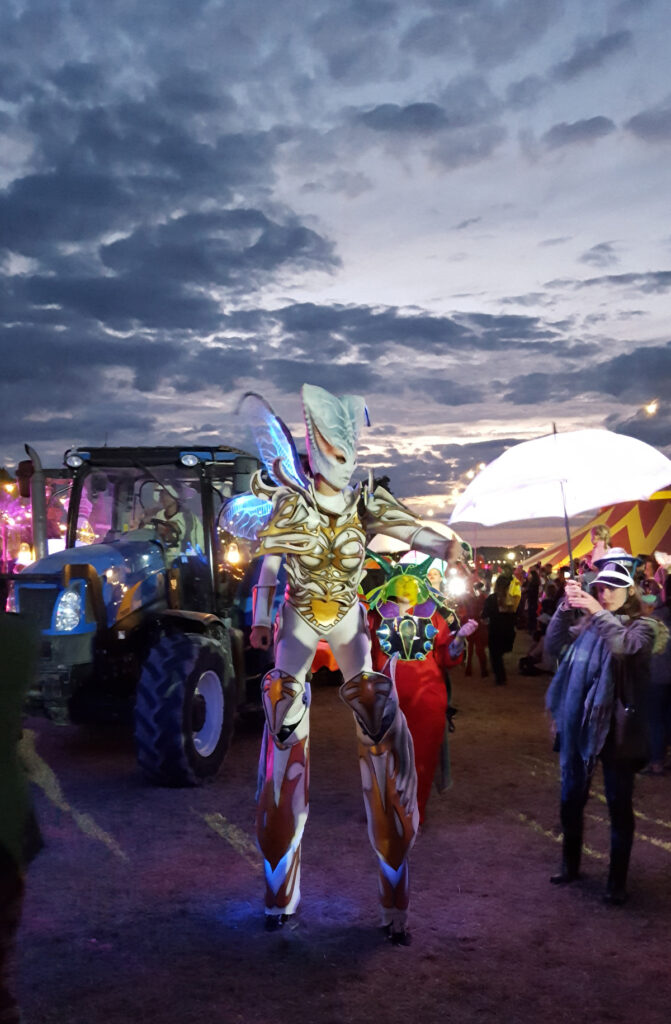 An actor dressed as an exotic alien, with glowing fairy wings and an exoskeleton that incorporates stilts, walks among the crowd at dusk