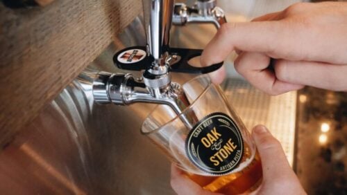 This image features a close-up of a person's hand pulling a beer tap to pour beer into a glass. The glass is branded with "Oak & Stone," which suggests it might be from a bar or a brewery of that name. The tap also has a label, although it's not fully legible in the photo. The focus is on the action of pouring the beer, with the beer itself appearing golden and foamy, indicating it is likely freshly poured. The background is out of focus, emphasizing the pouring action and the beer tap apparatus.
