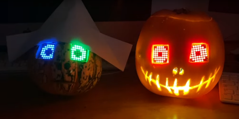 Two pumpkins on a desk in a darkened room with their LED eyes glowing in different colours. The LED matrices make the mouth on one pumpkin glow too