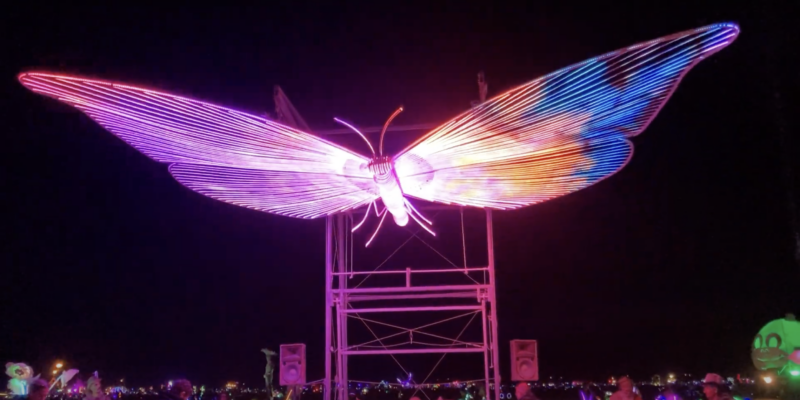 A giant butterfly currently glowing pink against a black sky. You can see people on the ground underneath it, looking tiny compared to the 26 foot width of the scuplture. Some scaffolding is visible behind the butterfly scultpure, presumably holding it 15 feet in the air