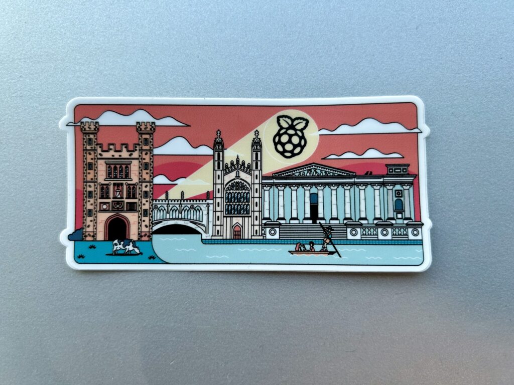 Our bat signal sticker featuring famous Cambridge buildings on the river against a sunset sky with a giant beam of light featuring the raspberry pi logo striking across the sky