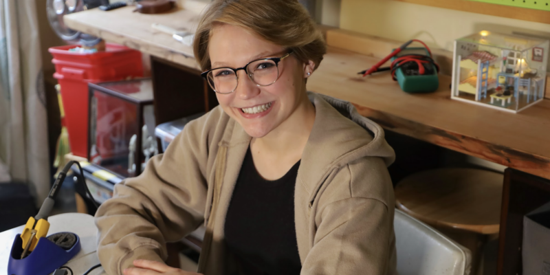 allie weber sat at a desk in a brown hoodie and black t shirt smiling at the camera which is held slightly higher than her head and off to the right