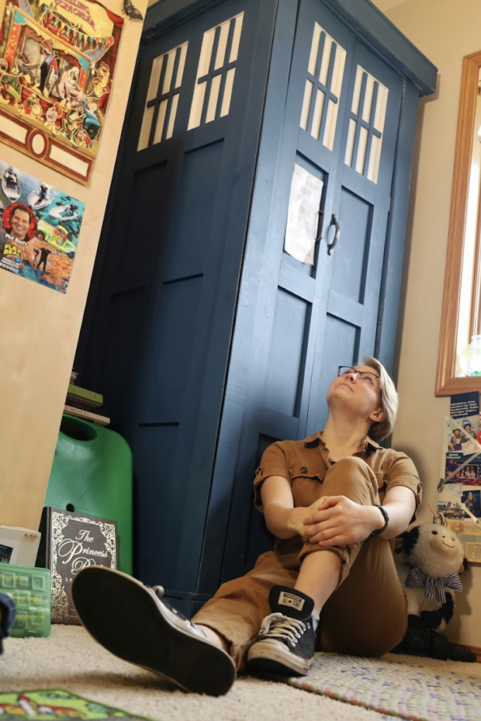allie weber wearing a brown boiler suit sat on the floor against a replica police box from doctor who