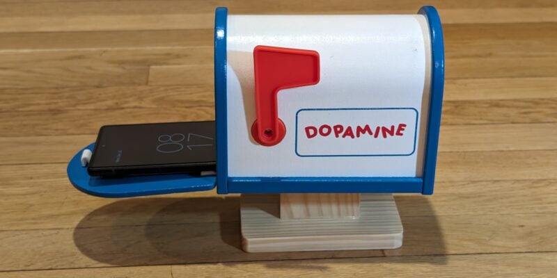 a tiny american style mailbox in white with blue edges and a red flag. the word dopamine is written on the side in red. it is sat on a wooden table