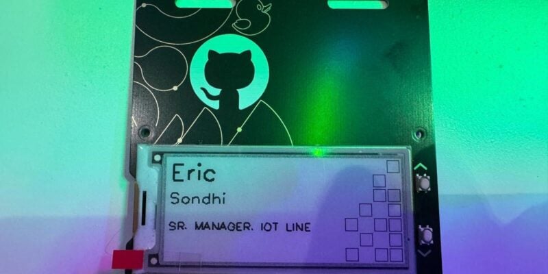 the front of the badge which is black and half taken up by a small screen with the name Eric written on it. On the black part of the badge with no screen, there is a white Git Hub logo