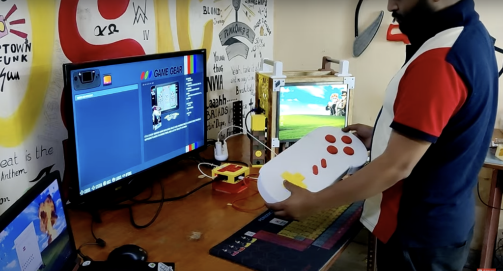 arnov sharma wearing a blue red and white polo shirt stood in front of a wooden desk with a PC screen on it running a game emulation desktop interface. He is holding a gaming controller the size of his torso with two hands.