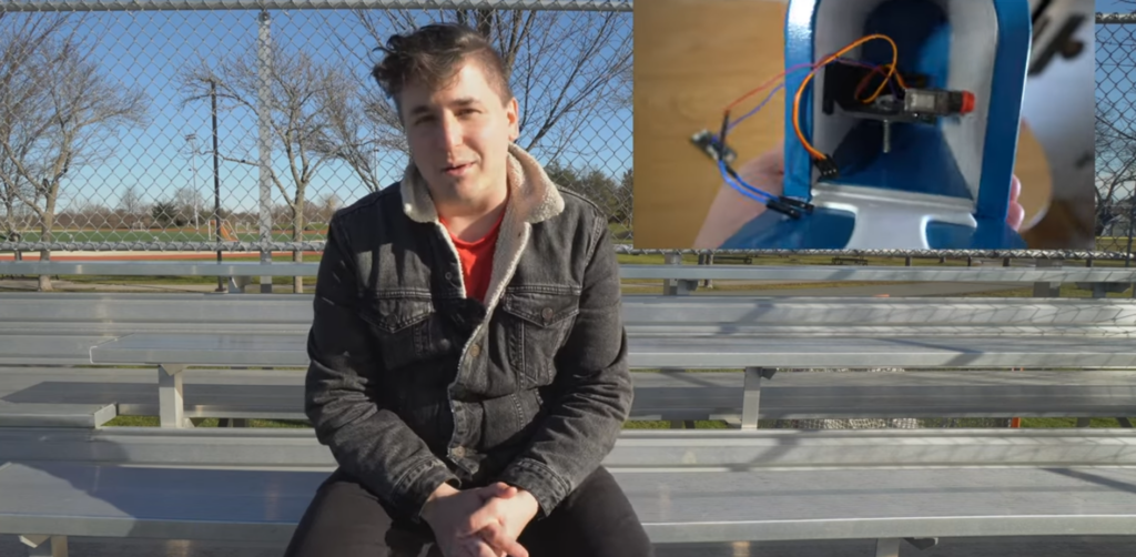 guy sat outside in a skatepark wearing a coat and talking about his creation. a break out window in the top right of the screen shows the inside of the mailbox with the hardware all wired in