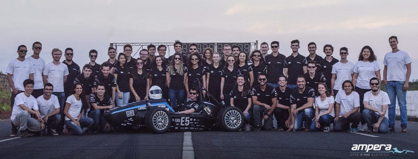 team ampera posing all together on a race track. most people are wearing matching black uniforms and the smaller groups on the left and right edges of the larger group in the middle are wearing light blue and white uniforms