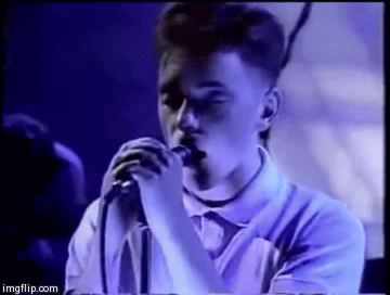 the lead singer of new order singing close into a microphone with the stage lights casting blue across him and his white polo shirt - he is singing blue monday