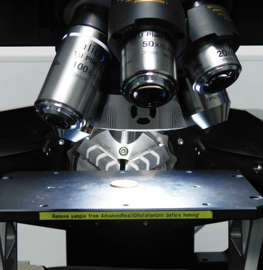 The image shows a microscope with three metallic cylindrical lenses labeled as “10x,” “50x,” and “20x.” These lenses are part of a revolving nosepiece for easy switching. Below the lenses, there is a mechanical stage where samples are placed for observation. The stage has holes and screws. A small circular sample is present on the stage. The background is dark, emphasizing the microscope and the sample. Text at the bottom warns to remove the sample from AdvancedReal3DRotationUnit before homing