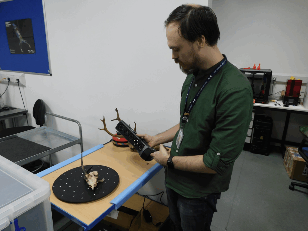 This image shows a 3D visualisation specialist at the Natural History Museum in London, who is scanning an object that looks like animal antlers with a skull fragment. The specialist is using a handheld device to control a laser scanner that is attached to a table.
