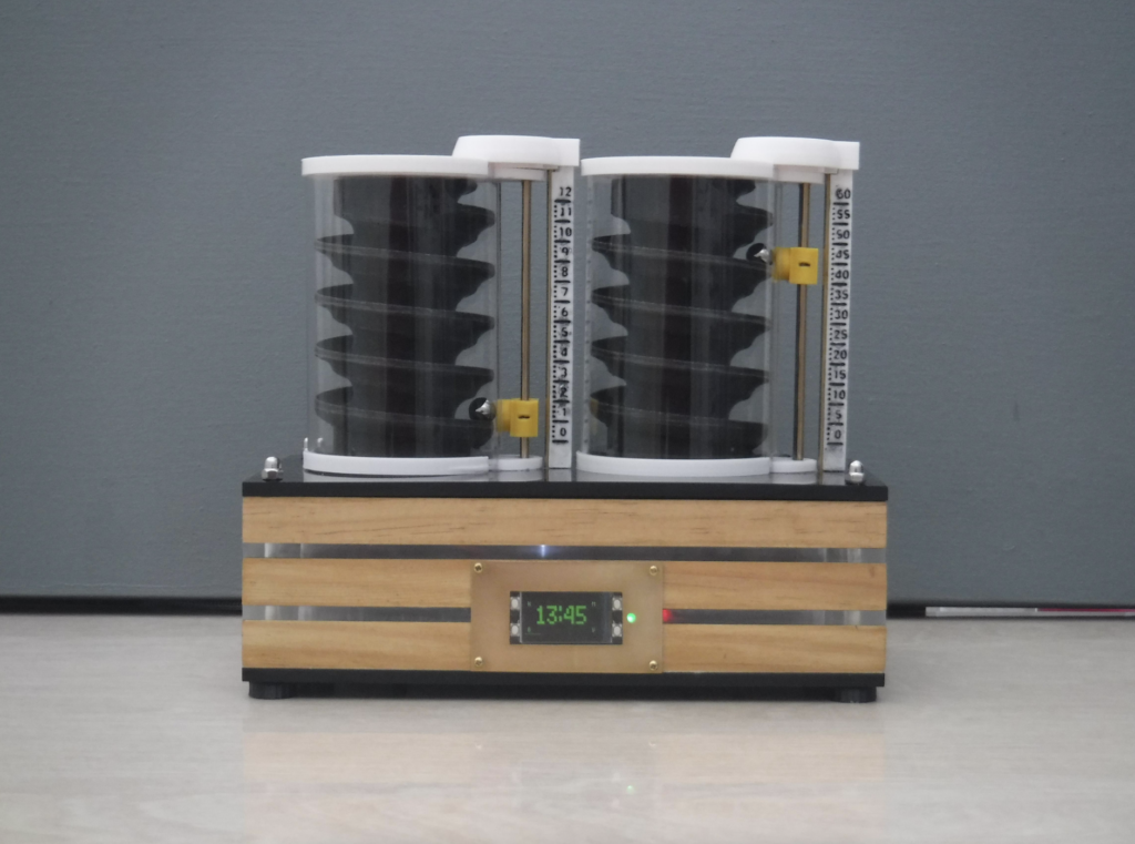 two cylindrical objects with spiral interiors, placed side by side on a wooden platform with a digital display showing “13:45”. The clock uses ball bearings to indicate the time.