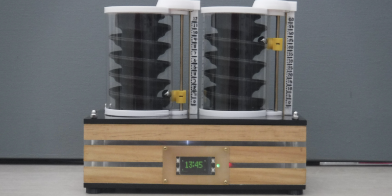 two cylindrical objects with spiral interiors, placed side by side on a wooden platform with a digital display showing “13:45”. The clock uses ball bearings to indicate the time.