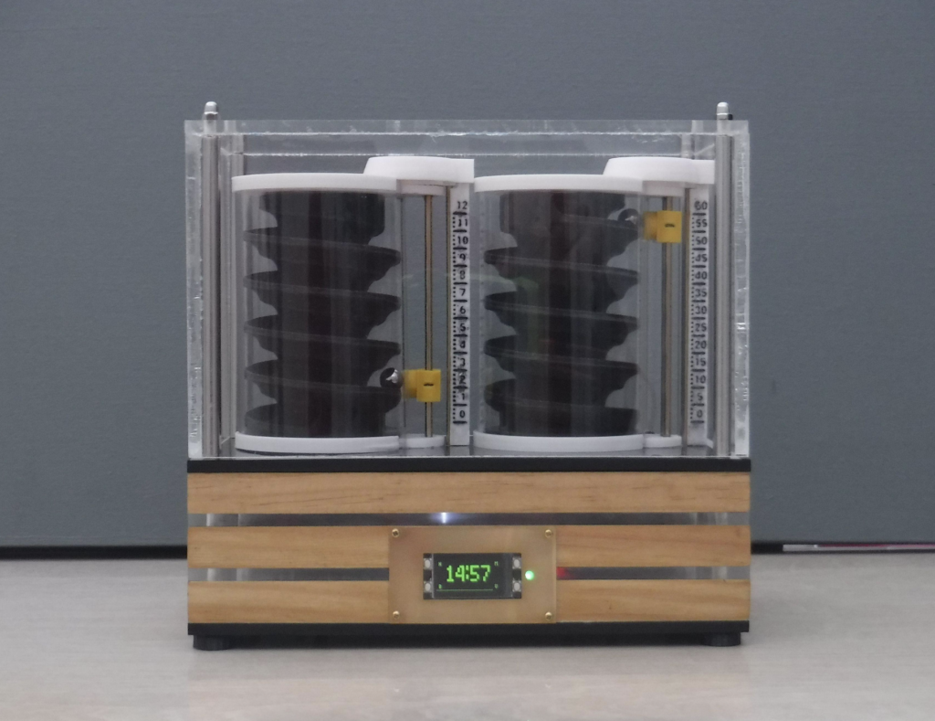 two cylindrical objects with spiral interiors, placed side by side on a wooden platform with a digital display showing “14:57”. The clock uses ball bearings to indicate the time.