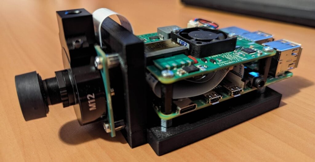 Device Components: The green circuit board hosts several electronic components, including connectors and ports.
Camera Module: A black cylindrical lens labeled “M12” is attached to one side of the device.
Cooling Fan: A small fan sits atop the circuit board, likely for cooling purposes.
Connectivity: Various USB ports and a ribbon cable are visible.
Sturdy Base: The assembly rests on a black base or stand.
Wooden Background: The device is placed on a wooden surface.