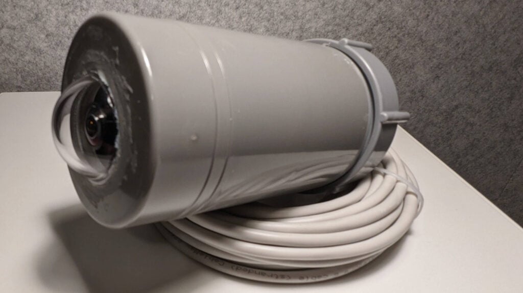 This image depicts a gray cylindrical camera with a visible lens at one end. The camera looks similar to those used for security or surveillance purposes. It shows signs of wear or residue on its body. A long white cable is coiled beneath the camera, likely for power and data transmission. The background features a textured gray wall and a flat surface where the camera rests.