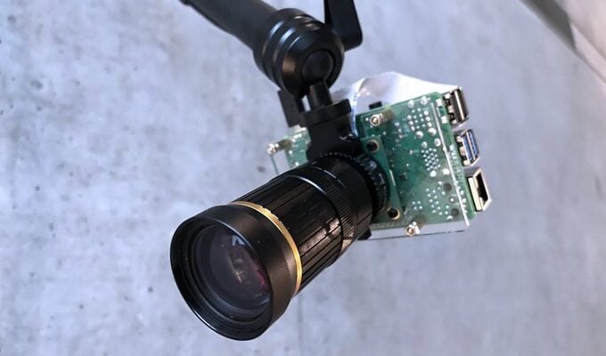 This photo depicts a camera module with an attached lens. The camera’s circuit board is exposed, and it’s mounted on some form of handle or stabilizing equipment. The large, prominent lens at the forefront has visible focusing rings and branding. The background is nondescript but appears to be a solid surface with some texture.