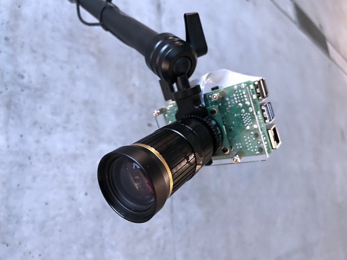This photo depicts a camera module with an attached lens. The camera’s circuit board is exposed, and it’s mounted on some form of handle or stabilizing equipment. The large, prominent lens at the forefront has visible focusing rings and branding. The background is nondescript but appears to be a solid surface with some texture.