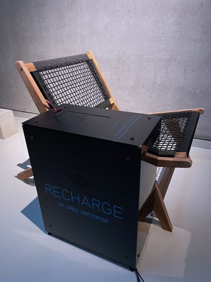 This photo portrays a modern, minimalist setting. A wooden chair with a black mesh seat and backrest is positioned next to a large black box labeled “RECHARGE by DRIES DEPOORTER.” The chair is made of light-colored wood, and the mesh material adds a contemporary touch. Against a grey concrete wall, the scene is illuminated by soft lighting from above. Visible cables and equipment enhance the industrial ambiance. The overall composition exudes relaxation and energy renewal.