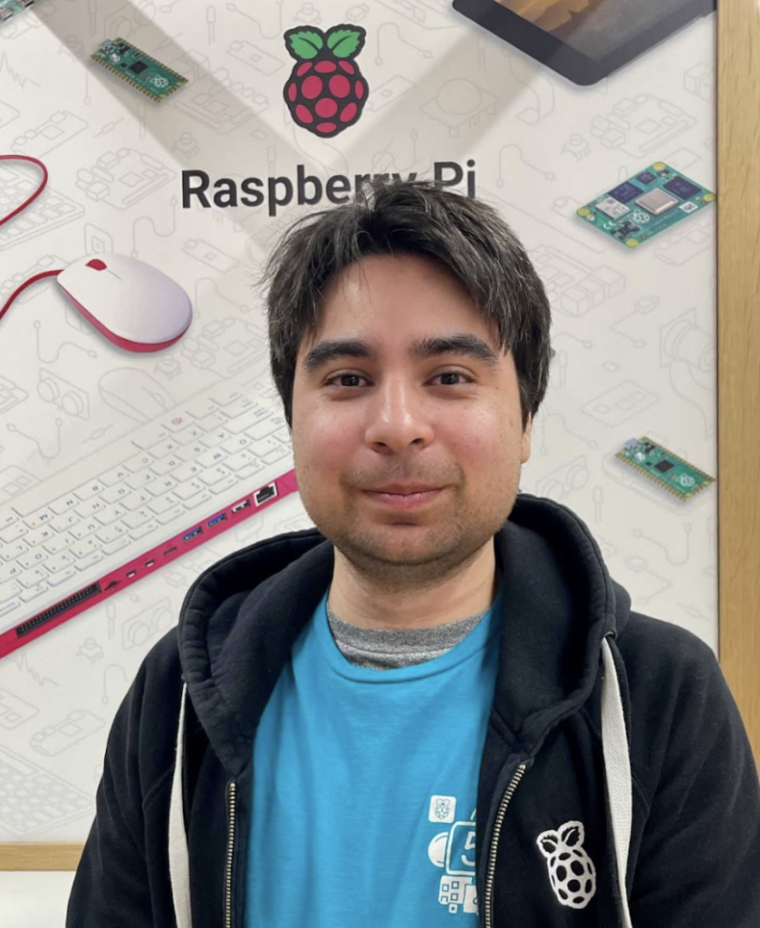tim stenning stood in front of graphics on the walls of the raspberry pi store. he is wearing a blue t shirt under a black hoodie. he has black hair and olive skin. he is looking straight into the camera