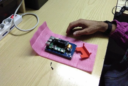 a person’s hand near an electronic circuit board on a pink anti-static mat. Here are the key details:

Circuit Board: The main focus is an electronic circuit board.
Human Element: A person’s hand, wearing a black watch, suggests work or examination.
3D Printed Object: An orange 3D printed object, likely related to the circuit board, is adjacent.
Workspace Setting: The setting indicates a workspace for assembling or repairing electronics.
