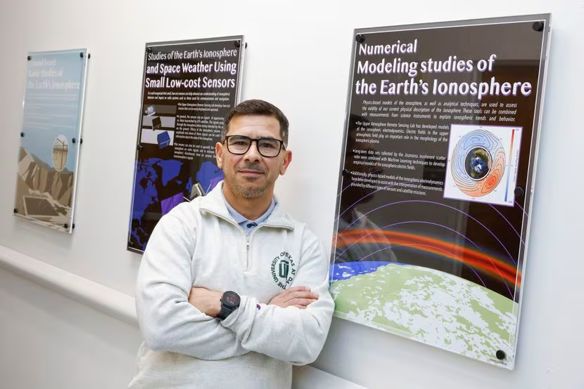 a person standing in front of two informational posters related to space science. Here’s a description based on the image details:

Individual’s Attire: The person is dressed in a white hoodie with an emblem and dark pants. Their face is not visible.
Poster Content:
The left poster is titled “Studies of Earth’s Ionosphere and Space Weather Using Small Low-cost Sensors,” which likely discusses research on the Earth’s ionosphere and the impact of space weather, using affordable sensor technology.
The right poster is titled “Numerical Modeling Studies of the Earth’s Ionosphere,” suggesting a focus on computational methods to study the ionosphere.
Both posters include text and visual elements such as graphs or data visualizations, and are displayed on a light-colored wall.