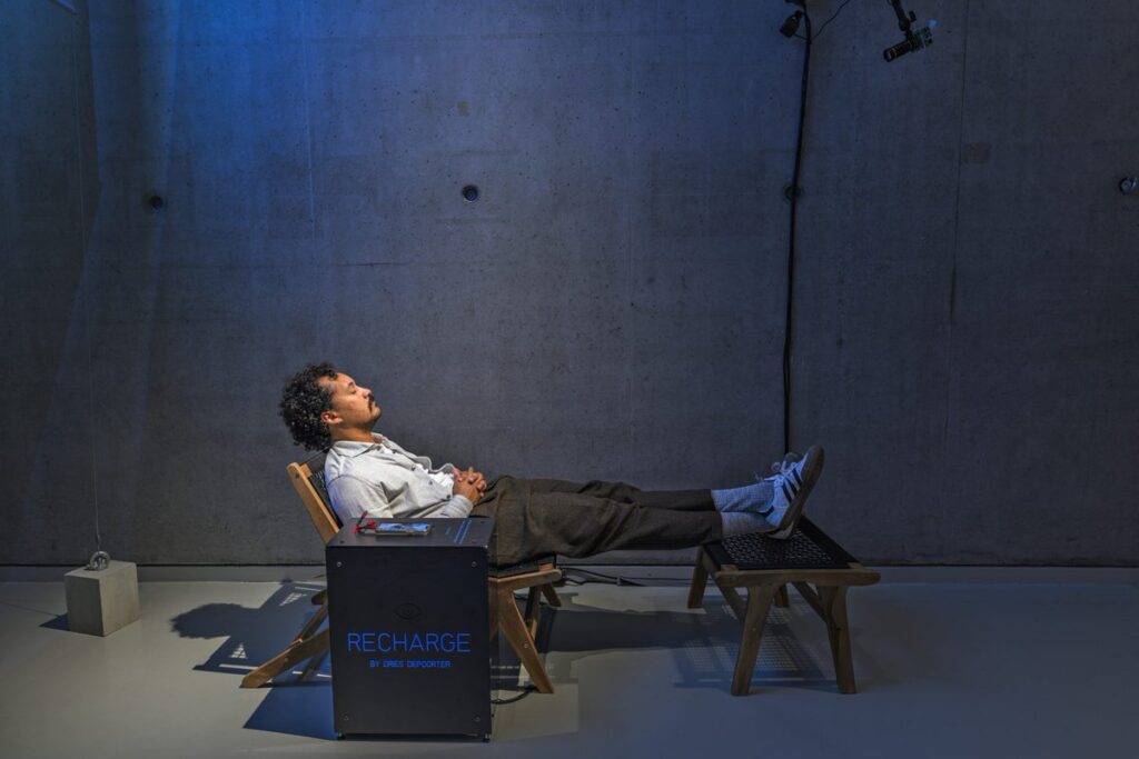 The image portrays a person reclining on a wooden chair with an extended footrest, appearing to be in a state of relaxation or recharging. The individual’s face is not visible, either blurred or covered for privacy. They are dressed in formal attire, including a white shirt and dark trousers. A box labeled “RECHARGE” sits beside the chair, suggesting the theme of relaxation or energy renewal. The setting is minimalistic, with a grey concrete wall in the background, illuminated by soft lighting from above. Some visible cables and equipment add an industrial touch to the overall ambiance.