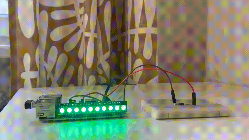 The image showcases a small electronic setup with a series of green LEDs that are illuminated. These LEDs are connected to a circuit board, which in turn is linked to a breadboard via red and black jumper wires. The focal point is a strip of green LEDs, brightly lit. It is attached to a small circuit board containing visible components and connectors. Two jumper wires—one red and one black—connect the circuit board to a white breadboard. The breadboard itself doesn’t have any other visible components. In the background, there’s a curtain with white and brown abstract patterns.