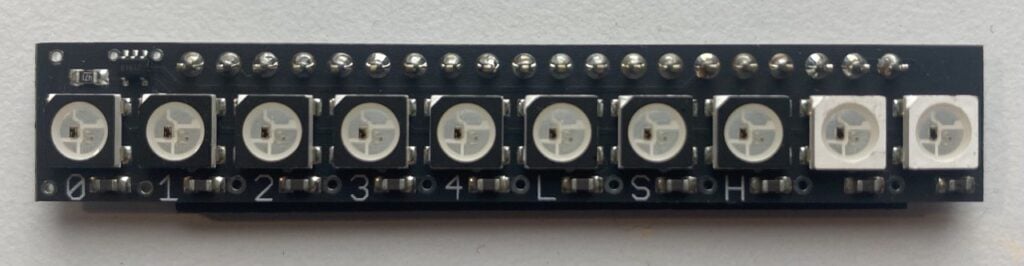 The image you provided shows a black circuit board with eight white LED lights mounted in a row. Each LED is encased in a square, white plastic housing. The circuit board has silver soldering points and connections visible. Numbers from 0 to 7 are printed on the board below each LED to indicate their positions. Unfortunately, the text and symbols at the left end of the circuit board are not clearly visible in the image. Overall, it appears to be a setup for controlling LED lights using a Raspberry Pi or similar hardware.