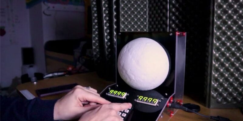 A screenshot from a DIY project video, showing a moon phase display prototype with LED lights illuminating a crescent moon model mounted on a rotating rod. The moon model casts a shadow onto a screen, simulating different moon phases as the rod spins. Various electronic components and wiring are visible in the background, indicating the technical complexity of the project.