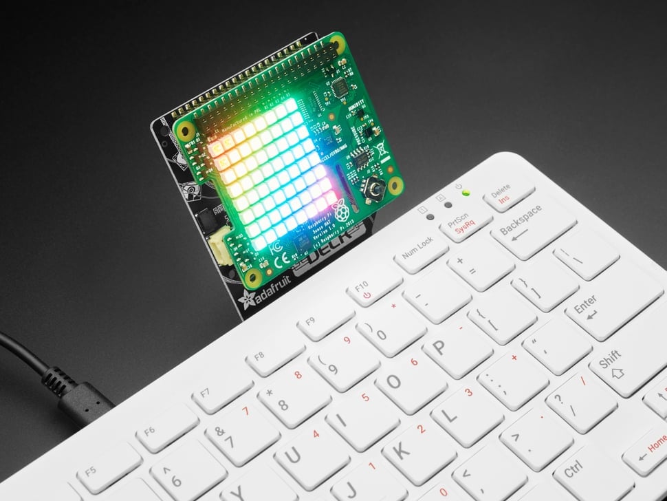 This image showcases an Adafruit LED matrix display connected to a white computer keyboard. Let’s break down the details:

LED Matrix Display:
The focal point is an Adafruit LED matrix that emits a vibrant gradient of colors.
The matrix is positioned in the upper right corner of the image.
It illuminates various hues, creating a visually striking effect.
Computer Keyboard:
The white keyboard occupies the majority of the image.
It features standard QWERTY keys, including special function keys like volume control, backspace, enter, shift, and control.
Four LEDs on the top right corner indicate different statuses; two of them are currently lit.
The keyboard rests on a dark grey or black surface, providing contrast against the colorful LED display.
Brand Mention:
The text “Adafruit” is visible, suggesting that this LED matrix display is likely an Adafruit product.
In summary, this image combines technology (the LED matrix) with a familiar input device (the keyboard), creating an intriguing juxtaposition of light and functionality.
