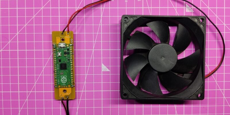 a Raspberry Pi computer module connected to a cooling fan, both laid out on a pink grid surface: The image shows a Raspberry Pi computer module and a black cooling fan placed on a pink surface with white grid lines. The Raspberry Pi is mounted on an elongated yellow circuit board with various electronic components visible. Red and black wires connect the Raspberry Pi to the cooling fan, indicating power supply or control connections. The cooling fan is black with seven blades and is encased in a square frame; it appears to be of standard design for electronic device cooling. The pink surface has white grid lines at regular intervals, resembling a cutting mat or workbench cover used in electronics or crafts
