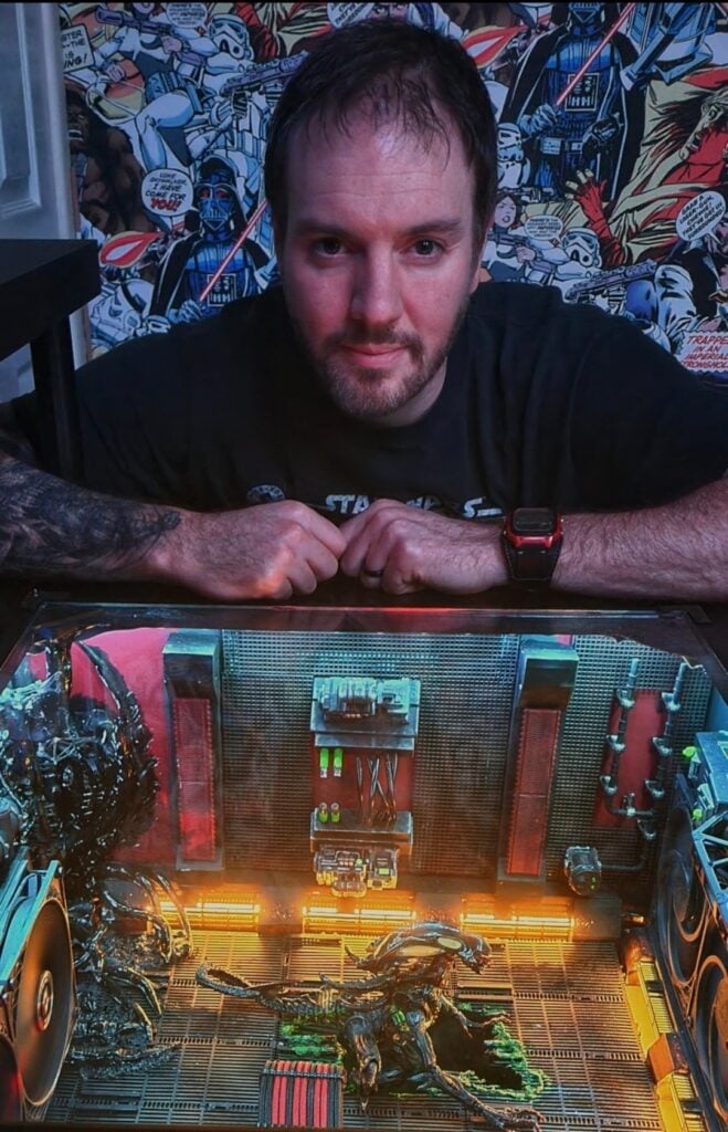 Custom-Built Computer:
The image features a person leaning over an open, custom-built computer case.
The individual’s face is obscured for privacy.
The person wears a black t-shirt and has tattoos on both arms.
The computer case is illuminated from within, showcasing its internal components.
Components include cooling fans, cables, and a decorative figurine resembling a dragon or mythical creature.
The background consists of colorful comic book pages covering the walls.
Artistic Aesthetics:
The overall mood combines technology and pop culture aesthetics.
The juxtaposition of the intricate computer setup against the comic book backdrop creates a visually striking scene