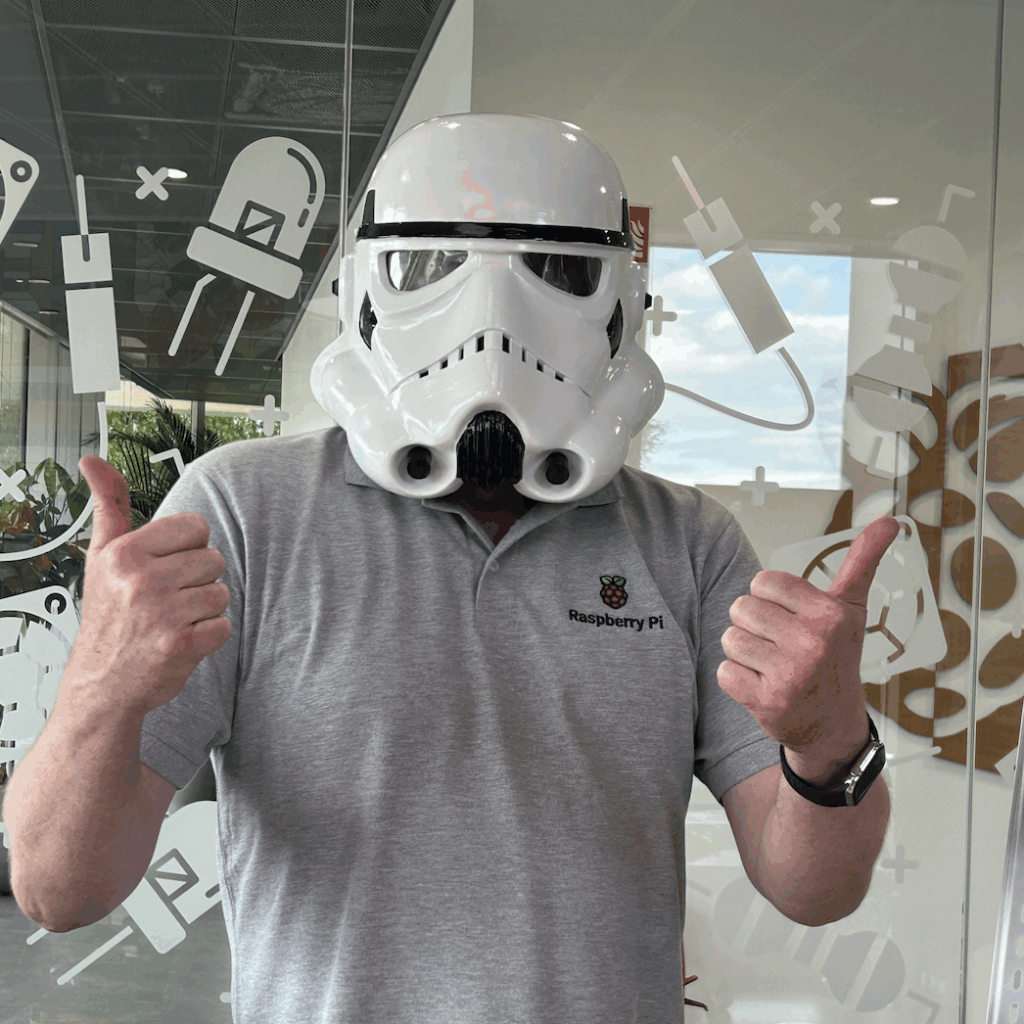 toby the maker wearing the helmet and a raspberry pi logo polo shirt standing in front of the decorated glass of the maker lab walls in pi towers