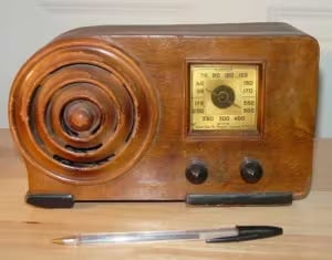 The image displays a vintage wooden radio from an earlier era, characterized by its classic design. The radio has a rounded, prominent speaker grille on the left, crafted with concentric circular patterns typical of mid-20th-century design. On the right side, there's a dial window displaying frequency ranges marked with both AM and FM bands, suggesting it's a multi-band receiver. Below the dial window are two black knobs, likely for volume control and tuning. To provide a sense of scale, there's a pen placed horizontally in front of the radio. The entire piece, with its wooden casing and design elements, exudes a nostalgic charm, reminiscent of the 1940s to 1950s radio design.