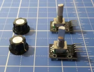 The image showcases a set of electronic components, specifically potentiometers, mounted on small breakout boards. There are three potentiometers in total. Two of them are equipped with black knobs, placed separately from the third component, which is mounted on a board and also features a silver metallic knob. The potentiometers on the breakout boards have pins on the bottom for easy integration with other electronic projects or breadboards. The setting is a blue grid background, typically used as a cutting mat or work surface in electronics or crafting, which helps in measuring and aligning components precisely. This setup is commonly used in DIY electronics for controlling volume, lighting, or other variables.