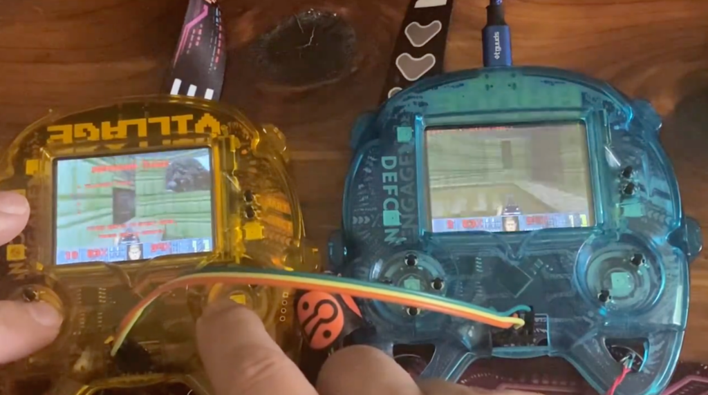 The image shows two handheld gaming devices, each running a classic first-person shooter game that appears to be "Doom." The devices are transparent, revealing their internal circuitry, and are connected with a multi-colored cable. The device on the left is yellow, and the one on the right is blue. The yellow device has "VILLAGE" written at the top, while the blue device has "DEFCON" and "ENGAGE" written on it. The screen of each device displays the game in progress, with visible health, ammo indicators, and the classic "Doom" heads-up display (HUD). The hands of a person are seen holding the yellow device, interacting with its controls. The devices are resting on a wooden surface, and there are lanyards attached to them with various designs, including one with hearts. The overall setup suggests that this is part of a custom or experimental gaming setup, possibly related to a tech conference or event.