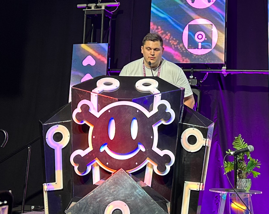 The image shows a man standing behind a large, decorated podium on a stage, likely giving a presentation or speech. The podium has a distinctive design featuring a large, illuminated smiling face with crossbones, reminiscent of a skull and crossbones but with a friendly expression. The design is highlighted by neon lights, giving it a vibrant and playful look. Behind the man, there are several screens displaying colorful graphics and icons that appear to be part of the event's theme. The stage is dimly lit, with the focus on the speaker and the podium. To the right of the podium, there's a small side table with a microphone and a potted plant, adding to the decor of the setup. The overall atmosphere suggests a tech or gaming conference, possibly with a hacker or cybersecurity theme.