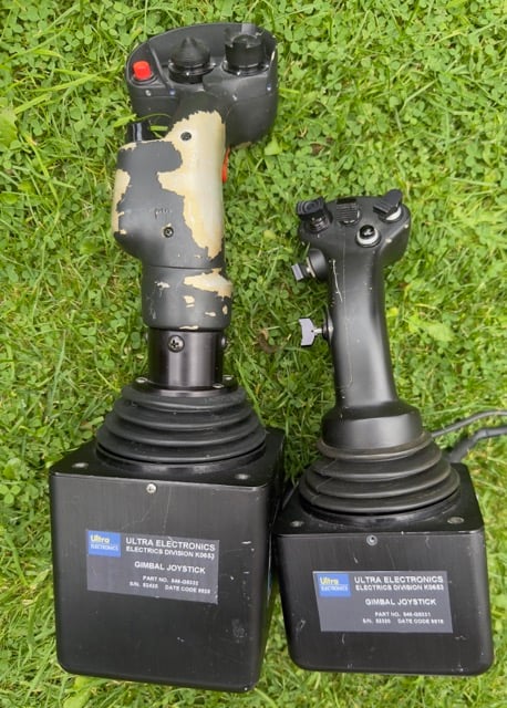 The image shows two gimbal joysticks resting on grass. These joysticks are industrial or military-grade, manufactured by "Ultra Electronics" (Electrical Division). They both have black bases with model information labels on them, identifying the joysticks as part of the Ultra Electronics line. Left Joystick: It appears worn, with patches of the original paint chipped away, revealing a lighter color underneath. The grip features several buttons and what looks like a control hat (small thumbstick). Right Joystick: This one appears to be in better condition, with a smoother and more consistent finish. It also has buttons and a control hat, similar to the left joystick, but the design is slightly more compact and less worn. Both joysticks are mounted on black gimbal bases, which allow for multidirectional movement, and each has a label with the Ultra Electronics branding. The labels contain text, including "Gimbal Joystick" and some part or batch numbers, but they are partially obscured or difficult to read due to the angle. These joysticks could be from fighter jets or advanced simulation setups.