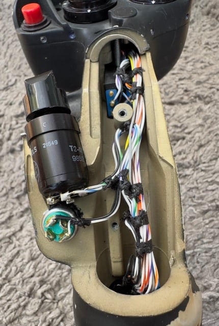 The image shows the internal view of a joystick, likely the same worn joystick seen in a previous image. The outer casing has been removed to reveal the internal wiring and components. Key elements include: Wiring: A bundle of color-coded wires runs vertically inside the joystick housing. The wires are neatly organized and secured with black ties, indicating they are part of the internal control system that connects the buttons and control mechanisms of the joystick to the external interface. Capacitor: A large black cylindrical component marked "2I649" and "T2" appears to be a capacitor. Capacitors like this are used for energy storage within electrical circuits and are common in joystick designs to manage power surges or smooth out signals. Metal Shaft: A metal rod or shaft is visible running vertically through the center of the joystick, likely part of the gimbal system that allows for multidirectional movement. Green Component: Near the bottom, there's a small green component with what looks like a circular interface or sensor, possibly related to the joystick's motion tracking or button functionality. Buttons: The top of the image shows part of the joystick grip with external buttons still visible, including a red button and a few other black controls. These buttons are connected to the internal components via the wiring. This internal view reveals the intricate electronic design and mechanical structure of the joystick, highlighting its complexity as a precision control device, likely for industrial or aviation purposes.