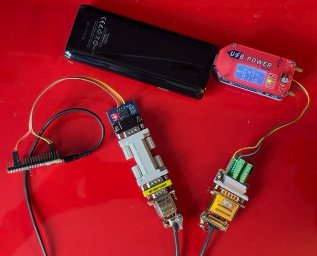 The image shows a collection of electronic components and connectors laid out on a red surface, likely part of a hardware setup for interfacing with or powering a device. Key components include: USB Power Supply: A black external battery pack is powering the setup via a USB cable. The USB cable is connected to a small red "USB Power" module with an integrated display showing output values, including 4.94V and 0.18A. This module is likely monitoring the power draw of the connected components. DB9 Connectors: There are two separate DB9 (serial) connectors. One has a grey body and a blue breakout board at the top, while the other appears to be connected to a green terminal block for wiring. These connectors are typically used for RS-232 or other serial communication interfaces, often found in industrial or retro computing hardware. Breakout Boards: The setup includes at least two small breakout boards attached to the serial connectors. These boards seem to have small ICs and wiring that enable communication or power regulation between the connected devices. Wiring: Several wires, color-coded in yellow, black, and red, are connecting various parts of the setup. Some wires are plugged into a pin header at the left, likely for GPIO or serial communication. Overall Setup: The combination of serial connectors, breakout boards, and USB power monitoring suggests this setup is being used for testing or interfacing with an external device, perhaps an older serial-controlled system or custom hardware. The USB power module ensures that the components receive regulated power, while the serial connectors enable communication between devices. This configuration looks like a development or testing setup for a project involving serial communication and low-power electronics.