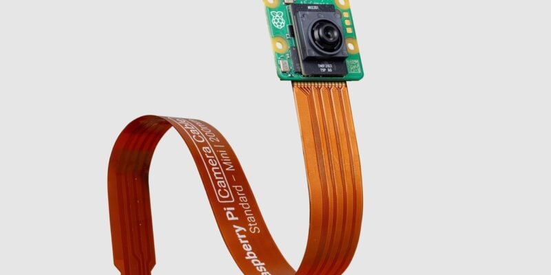 This image features a Raspberry Pi AI Camera Module connected to a long, curved orange ribbon cable. The small, square-shaped green circuit board has a black camera lens at its center and yellow mounting holes at each corner. The ribbon cable is flexed into a loop and prominently displays white text that reads "Raspberry Pi Camera Cable Standard – Mini – 200mm." The cable is designed to connect the camera to a Raspberry Pi device, and the image is set against a plain gray background.