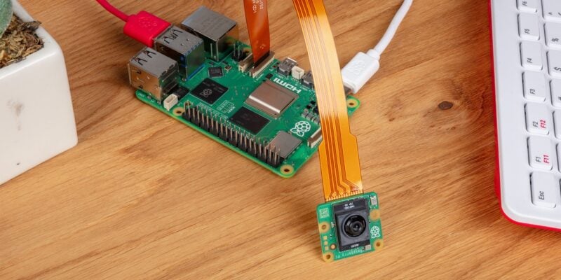This image shows a Raspberry Pi setup on a wooden surface, featuring a Raspberry Pi board connected to an AI camera module via an orange ribbon cable. The Raspberry Pi board is attached to several cables: a red one on the left for power and a white HDMI cable on the right. The camera module sits in the lower right corner, with its lens facing up. Part of a white and red keyboard is visible on the right side of the image, and a small plant in a white pot is partially visible on the left. The scene suggests a Raspberry Pi project setup in progress.