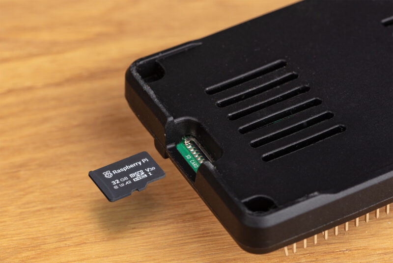 The image shows a microSD card partially inserted into a Raspberry Pi case. The card is branded with the Raspberry Pi logo and labeled as a 32 GB microSD card, which is commonly used to store the operating system and files for a Raspberry Pi device. The case has ventilation slots and appears to be designed to protect the Raspberry Pi while allowing easy access to the microSD card slot. The setup is likely intended for a compact and accessible way to operate and manage the Raspberry Pi.